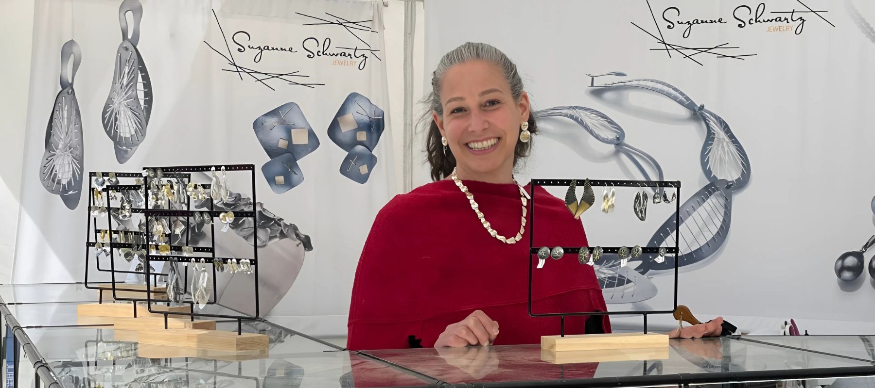 Suzanne Schwartz showcasing her unique jewelry designs at an exhibition booth, standing in front of a display featuring her modern, metal-stitched earrings and necklaces.